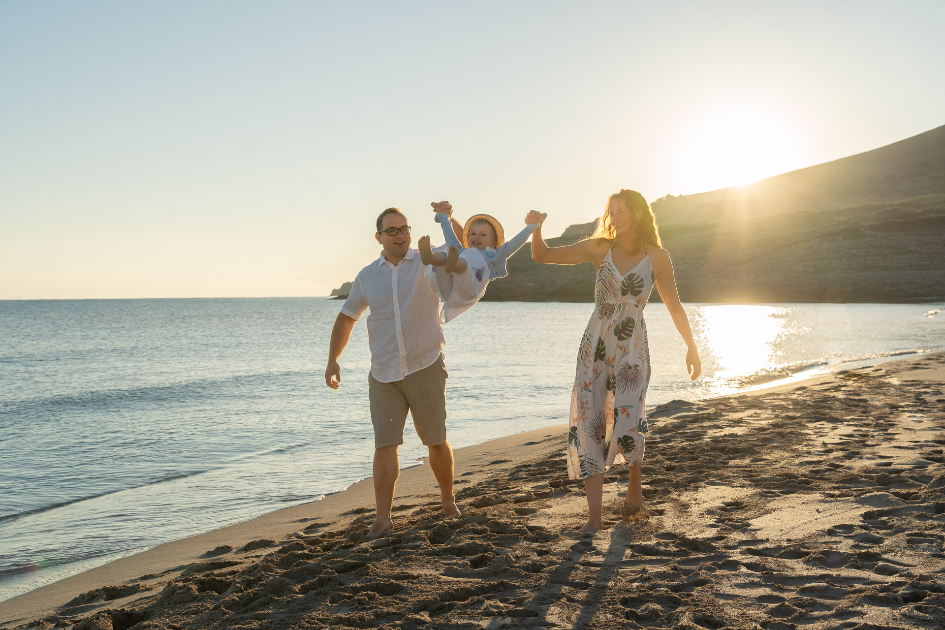 Cala Mesquida Familienfotoshooting – Eltern schwingen lachendes Kind am Sandstrand beim Sonnenuntergang auf Mallorca.