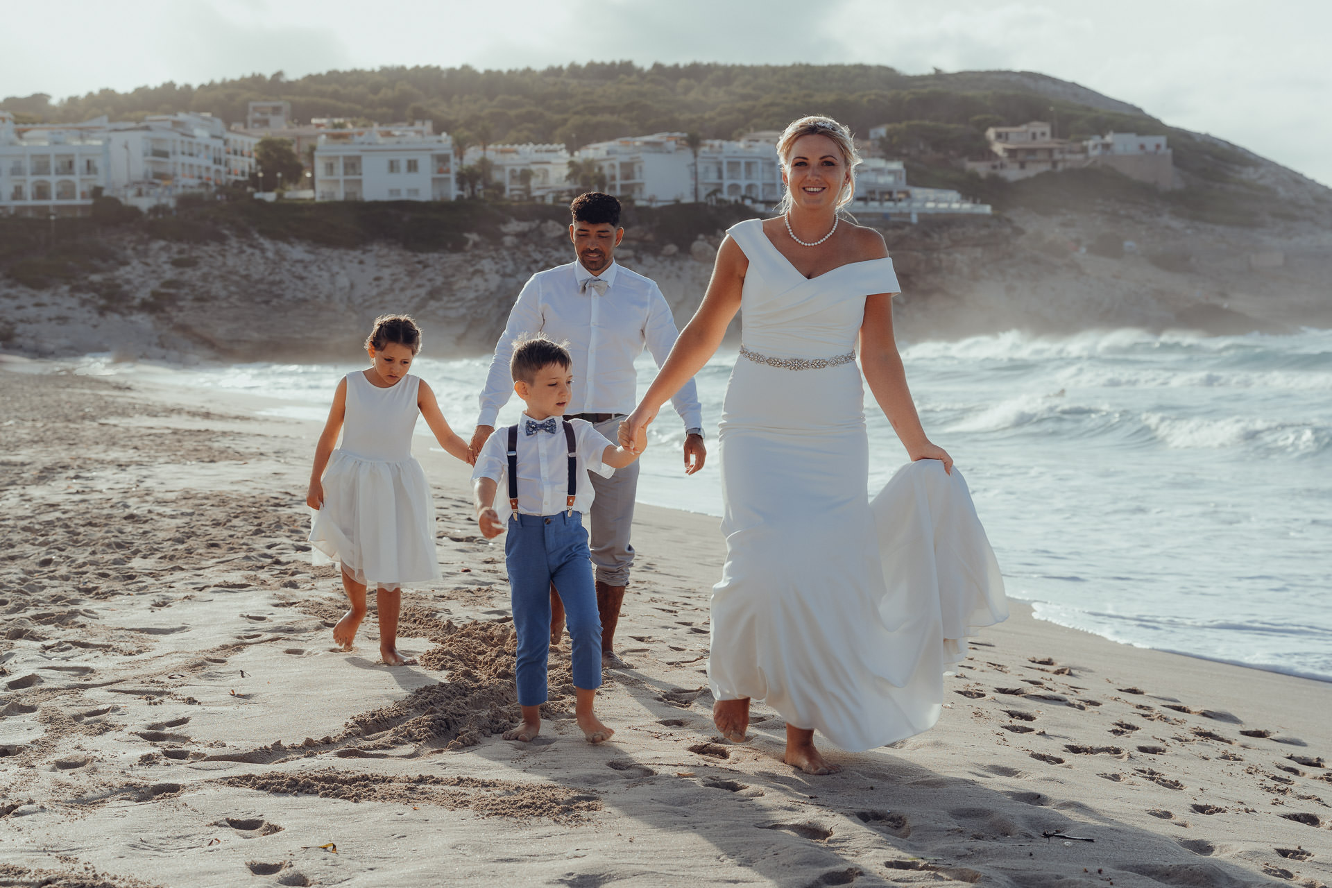 Cala Mesquida Strandhochzeit – Brautpaar mit Kindern läuft barfuß am Wasser, emotionale Hochzeitsfotos auf Mallorca.