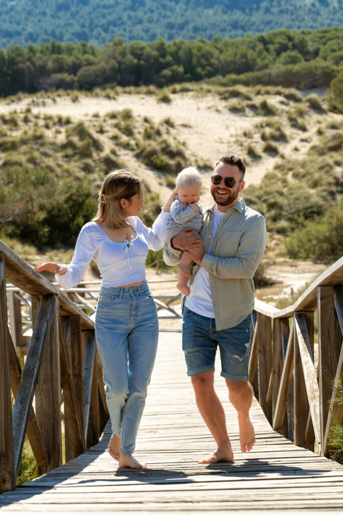 Junges Paar mit Baby auf dem Arm läuft über den Holzsteg an der Cala Mesquida