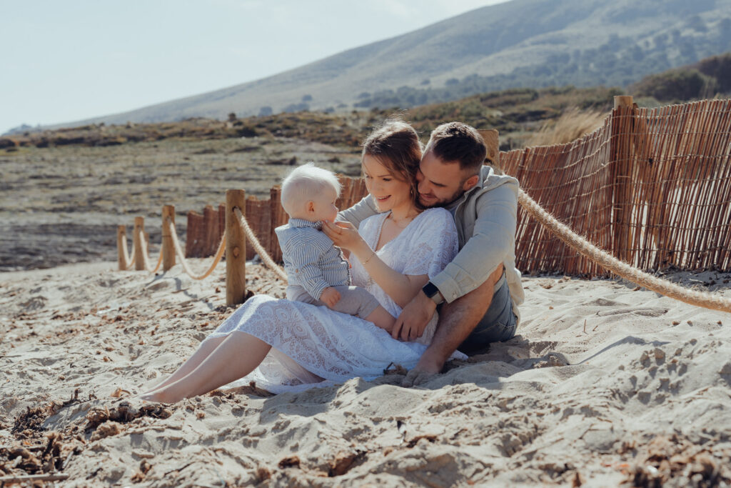Junges Paar sitzt beim Fotoshooting an der Cala Mesquida mit dem Baby im Sand