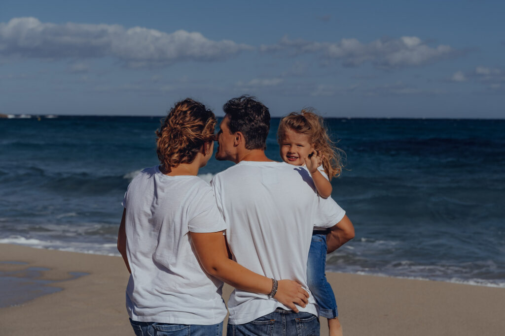 Kleine Familie steht am Meer der Cala Mesquida und schaut aufs Meer, während die kleine Tochter auf dem Arm des Vater in die Kamera lächelt.