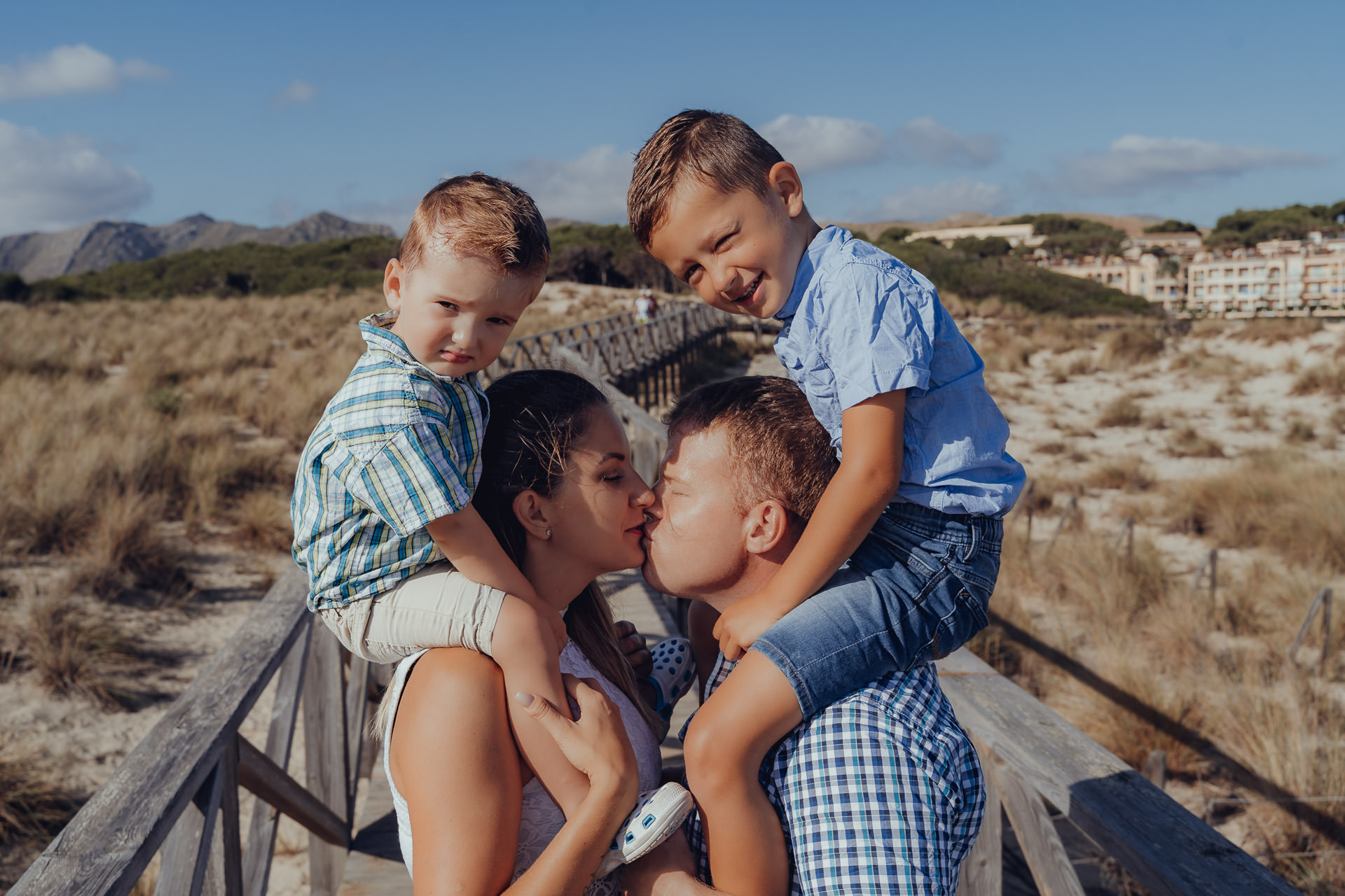 Familienfotoshooting Cala Mesquida – Eltern mit zwei Kindern auf den Schultern, Sommer, Dünen und Holzsteg auf Mallorca.
