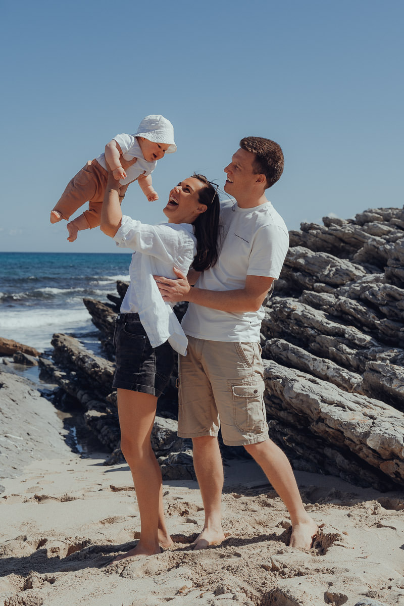 Cala Mesquida Fotoshooting Junge Eltern halten ihr Baby beim Fotoshooting hoch in den Himmel, im Hintergrund ist das Meer an der Cala Mesquida zu sehen.