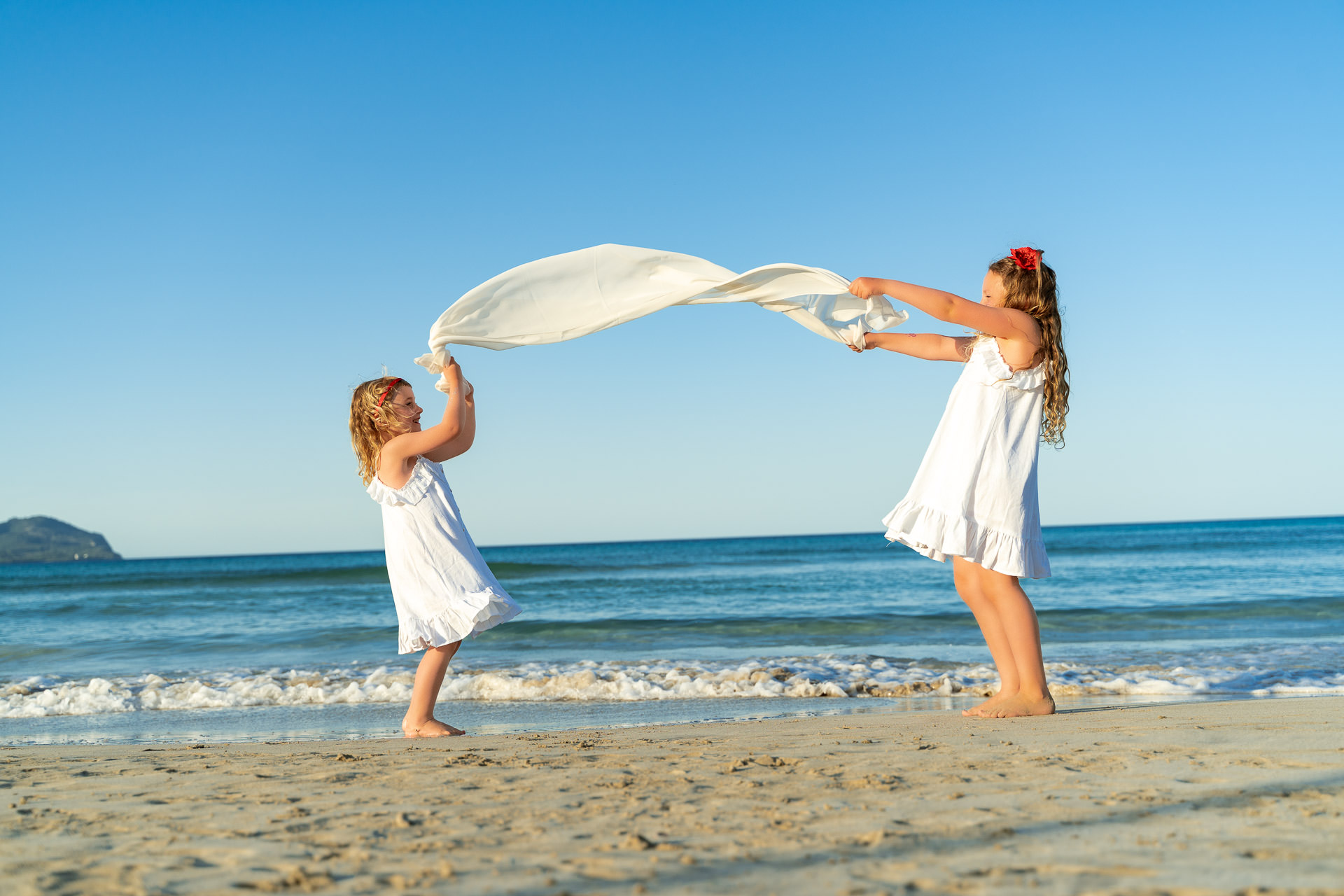 Zwei Mädchen spielen am Strand von Cala Millor mit einem weißen Tuch und lachen.
