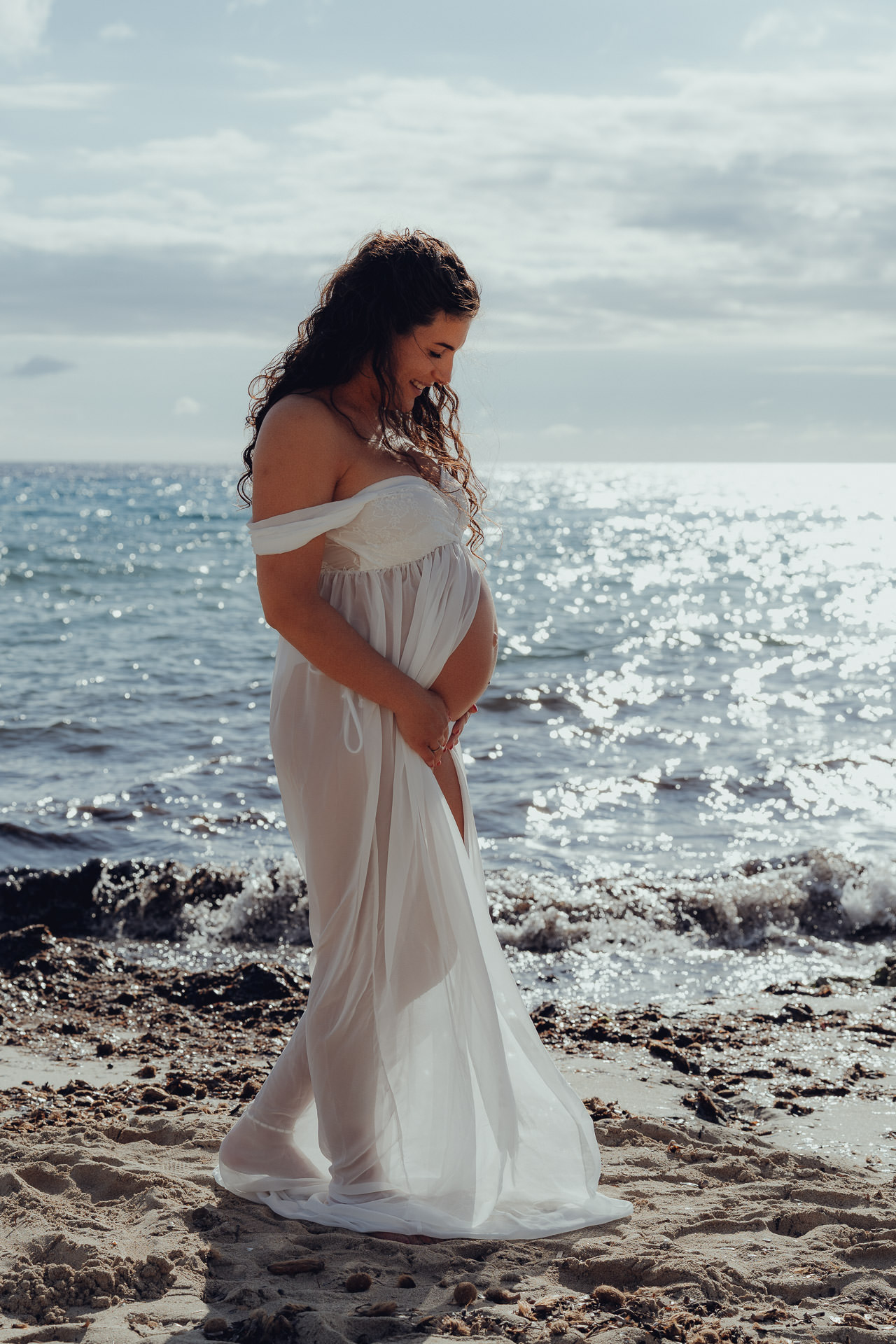 Schwangere Frau in weißem Kleid steht am Meer von Cala Millor und hält liebevoll ihren Babybauch.