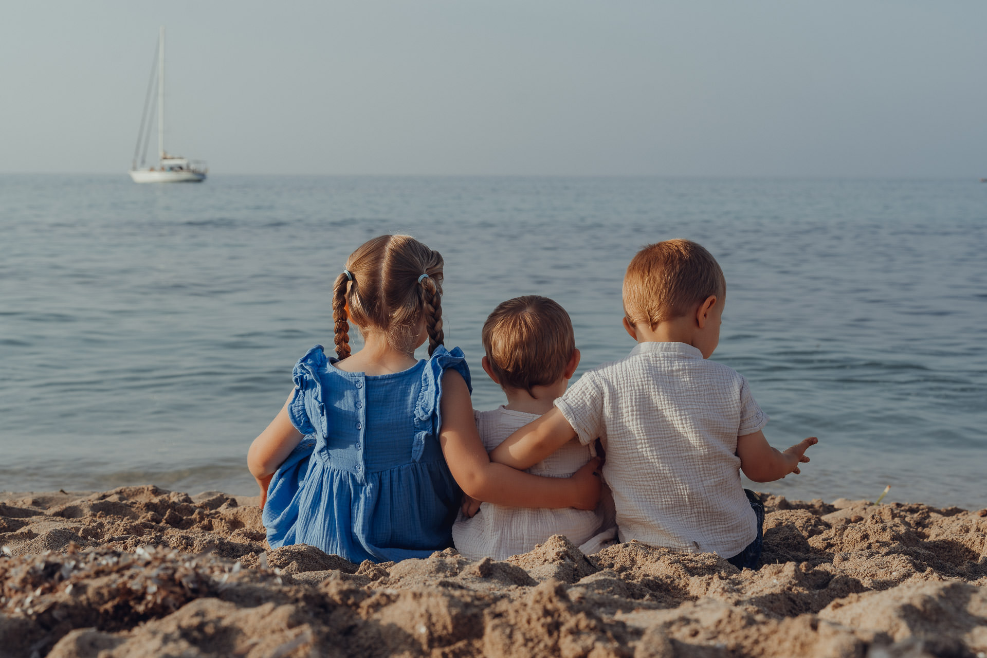 Cala Millor Fotoshooting Drei kleine Kinder sitzen Arm in Arm am Strand und schauen aufs Meer.