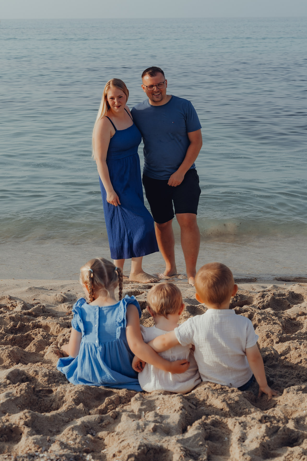 Cala Millor Fotoshooting Drei kleine Kinder sitzen am Strand Arm in Arm und schauen zu ihren Eltern die am Meer stehen.