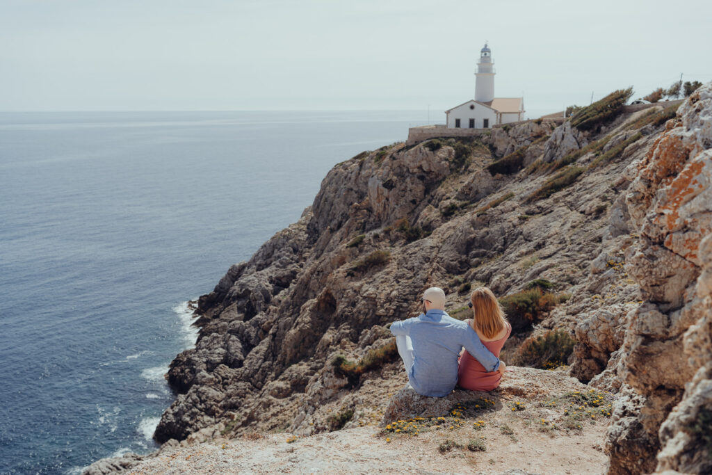 Junges verliebtes Pärchen sitzt auf den Felsen am Leuchtturm in Cala Radjada und blickt auf Meer
