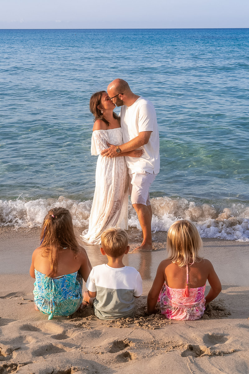 Cala Rajada Fotoshooting Drei Kinder sitzen im Sand und schauen zu den Eltern die sich küssen und mit den Füßen im Meer stehen
