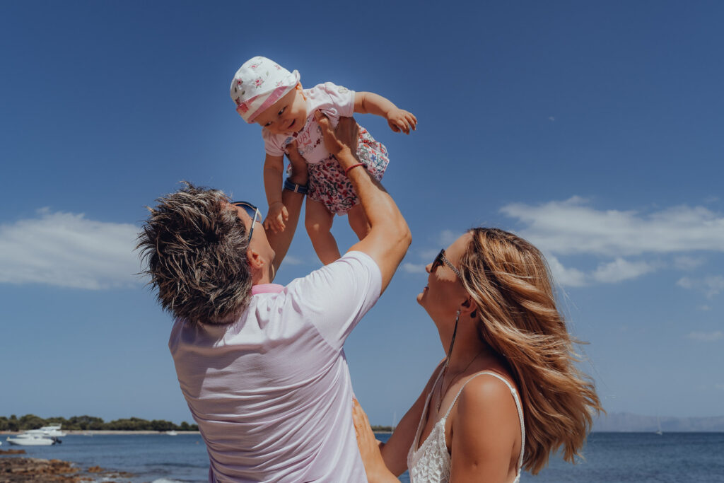 Papa hebt Baby lachend in die Luft während Mama zuschaut am Strand von Can Picafort