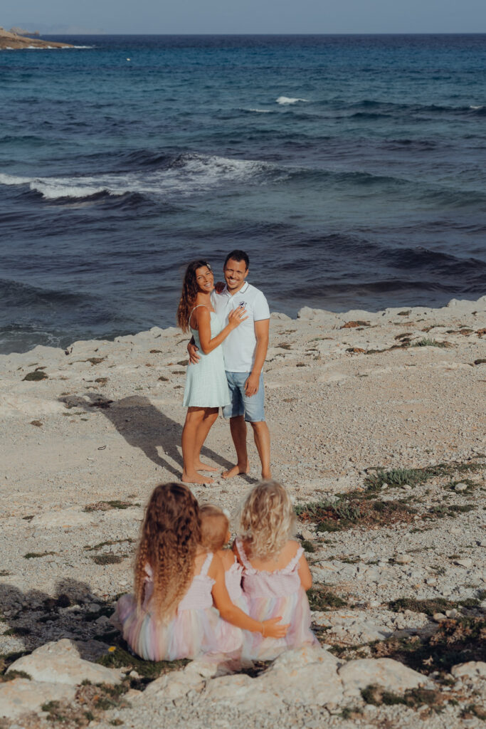 Eltern stehen mit drei Kindern am Felsen und blicken auf das Meer in Can Picafort