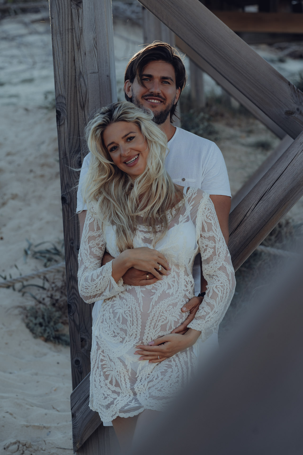 Elegantes Babybauch Shooting auf Mallorca – schwangeres Paar in Spitze unter Holzsteg am Sandstrand.