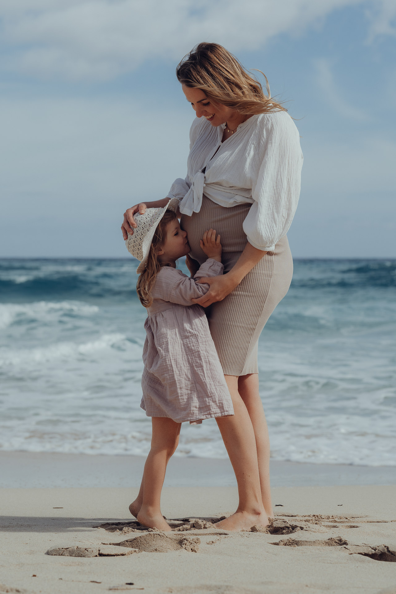 Natürliches Babybauch Shooting auf Mallorca – Tochter küsst liebevoll den Babybauch ihrer Mutter am Meer.