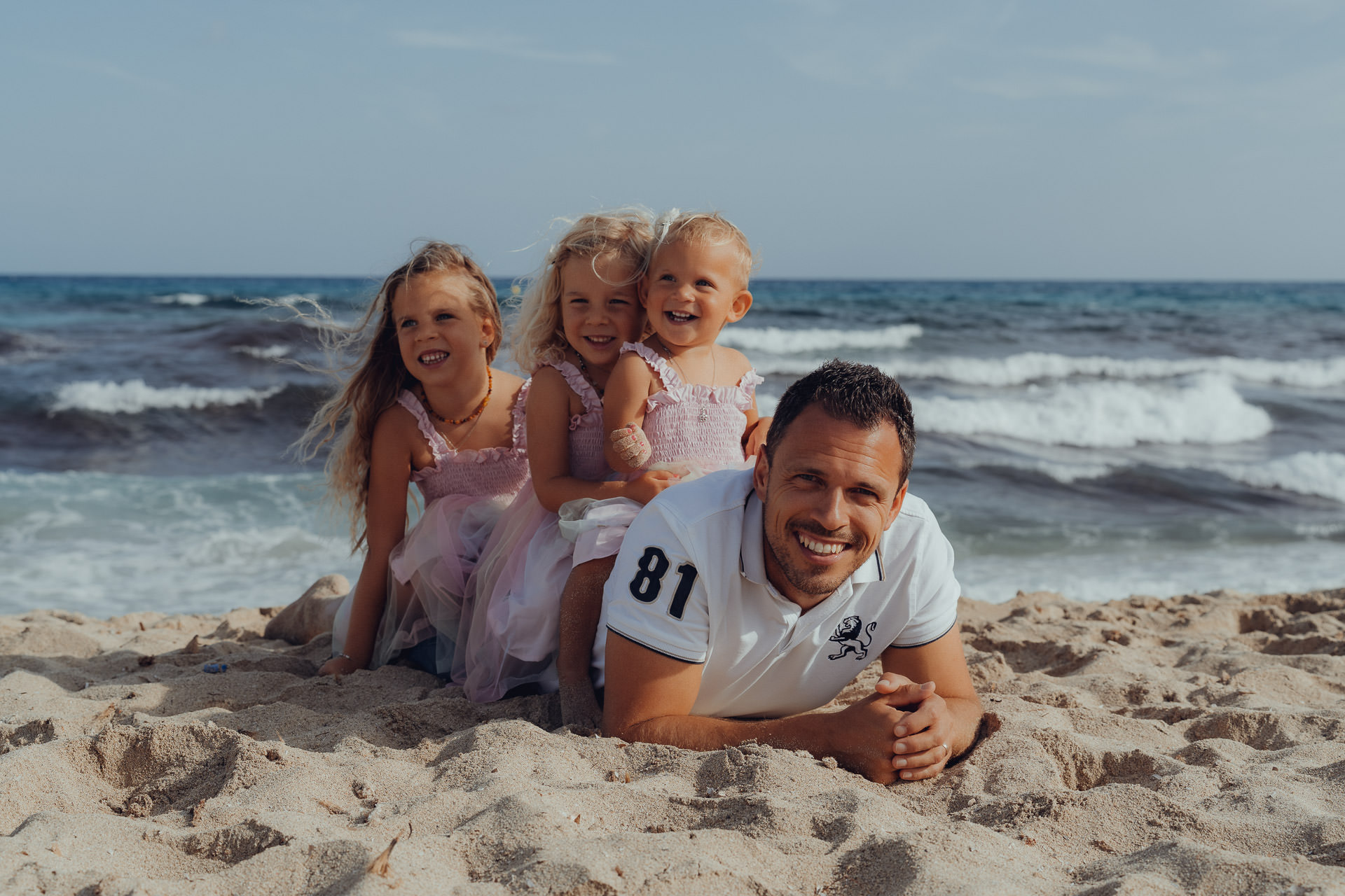 Vater liegt am Strand von Mallorca, drei kleine Töchter sitzen auf seinem Rücken und lachen