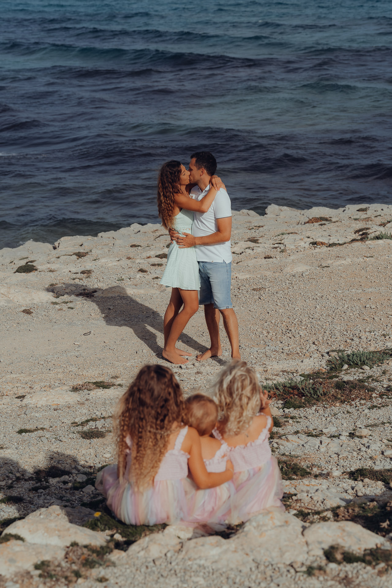 Eltern küssen sich am Strand von Mallorca, kleines Kind spielt unscharf im Vordergrund