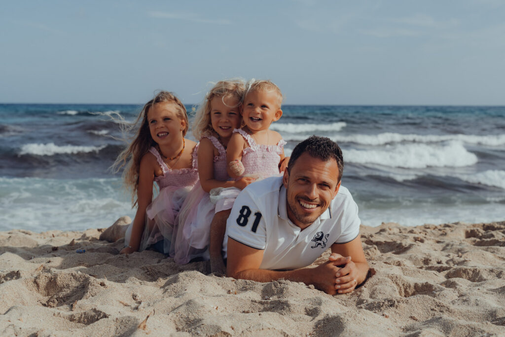 Drei kleine Mädchen sitzen auf dem Rücken vom Pappa der am Strand von Cala Millor liegt.