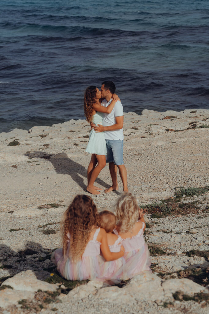 Drei kleine Geschwister sitzen an der Cala Mesquida auf den Felsen und schauen zu den Eltern die siich küssen