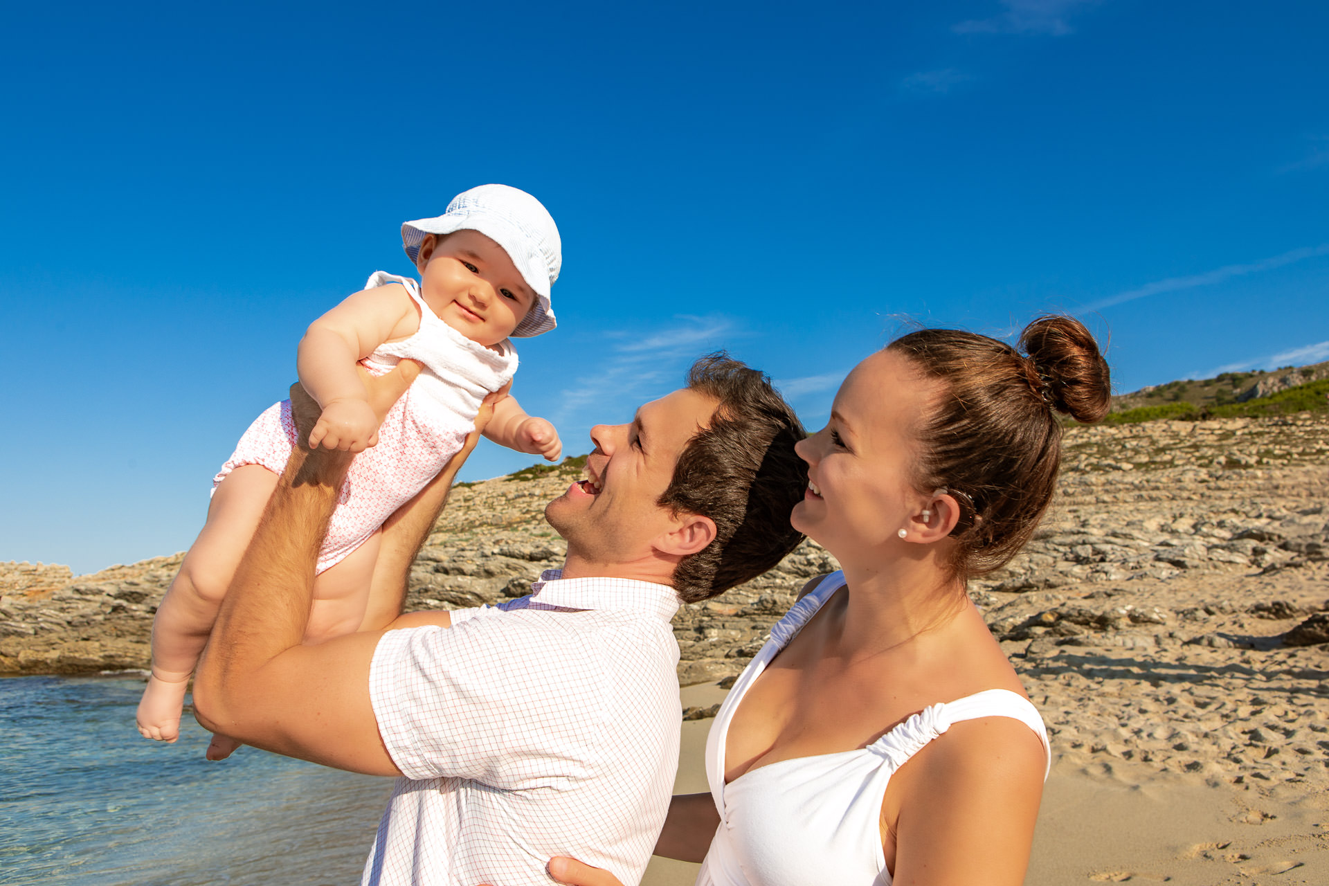 Vater hält Baby in den Armen, Mutter lacht, Familienglück am Strand von Mallorca