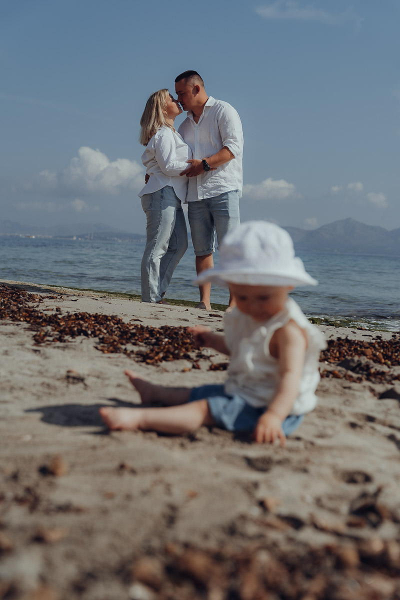 Mallorca Familien Shooting Baby sitzt unscharft im Vordergrund und die Eltern stehen im Hintergrund und küssen sich am Strand