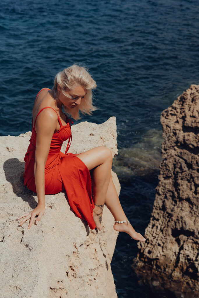 Model im roten sexy Kleid sitzt auf dem Felsen am Meer