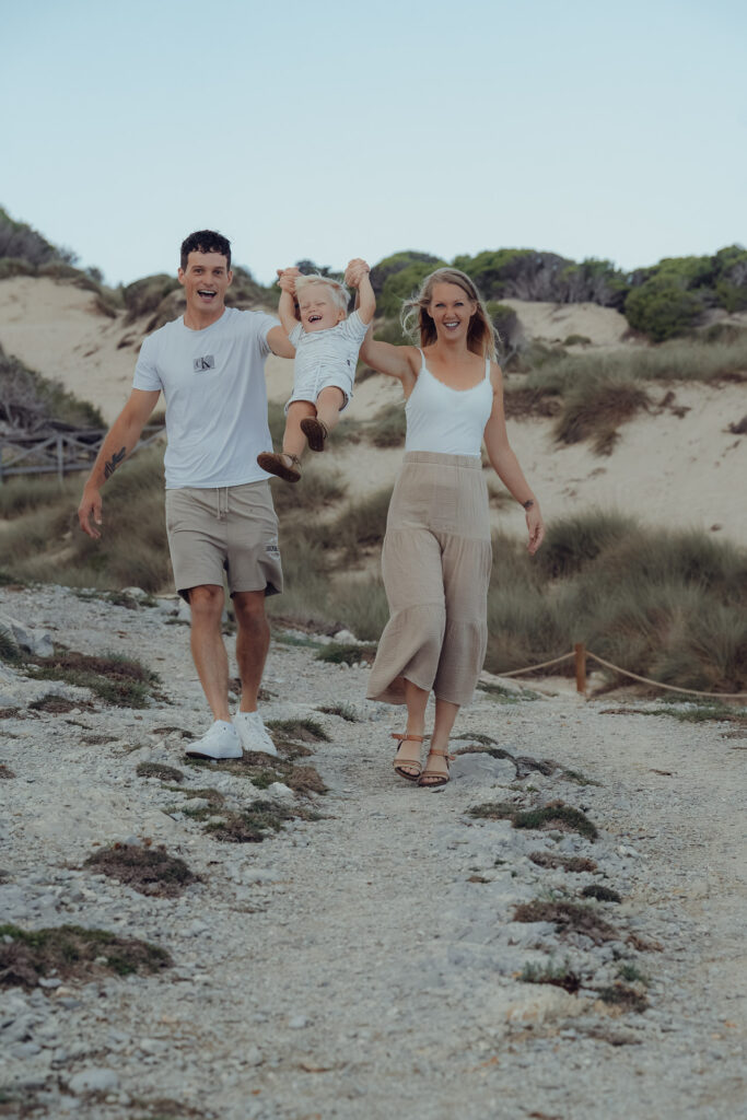 Lebendige Familienfotos in den Dünen Mallorcas – Eltern schwingen ihr Kind lachend zwischen sich. Mallorca Fotograf für natürliche Urlaubserinnerungen.