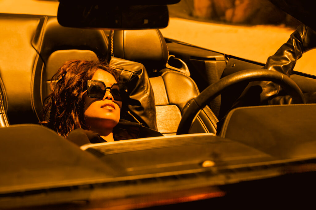 Dunkel Haariges Asiatisches Model sitzt lässig mit Sonnenbrille in ihren offenen Cabrio und genießt den Sonnenuntergang