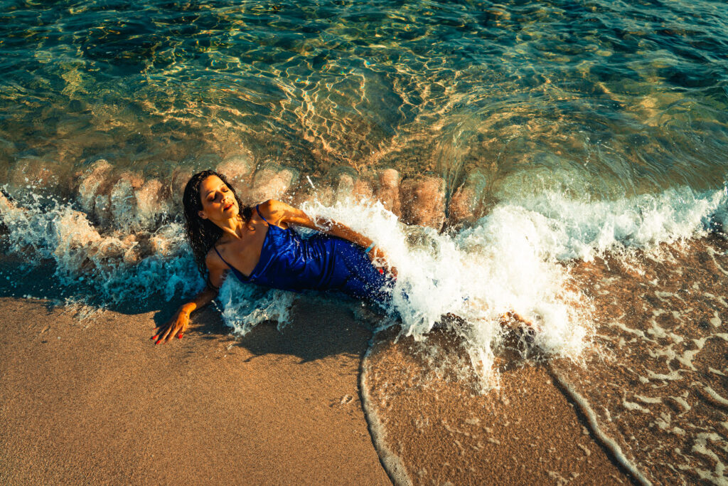 Model liegt mit blauem Kleid am Strand der Cala Mesquida während die Welle von hinten sie überrollt.