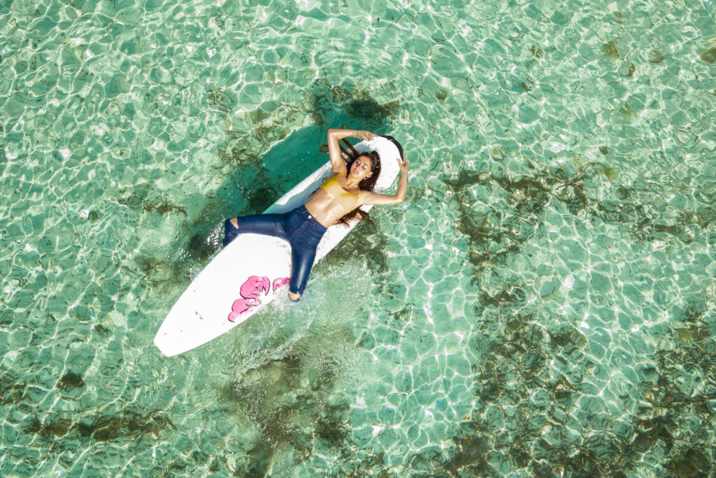Model liegt auf einem Surfbrett in nasser enger Jeanshose und Bikini Oberteil, das Bild wurde mit einer Drohne von oben aufgenommen.
