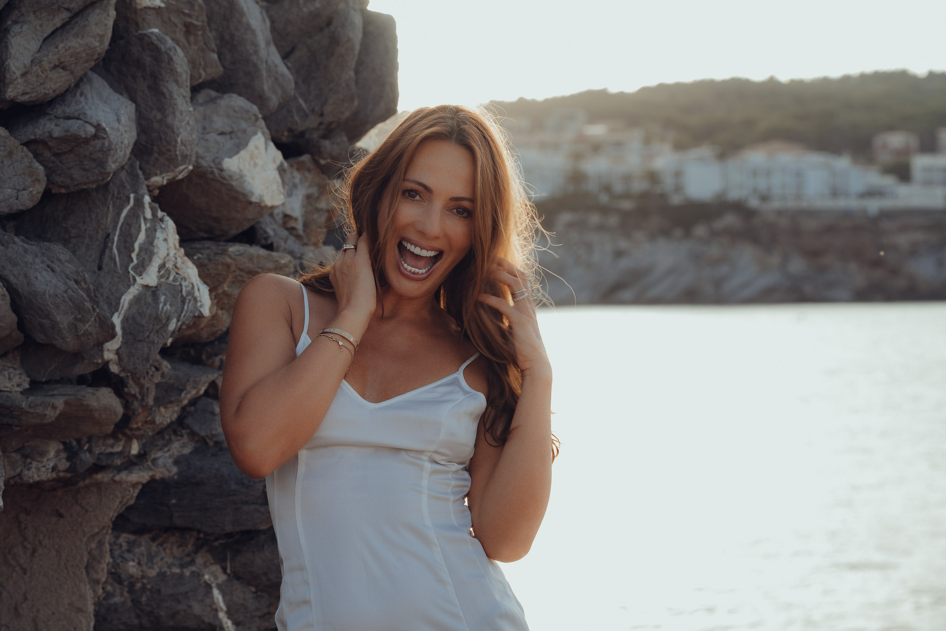 Lachendes Model im weißen Sommerkleid an der felsigen Küste, natürliches Mallorca Fashion Shooting am Meer
