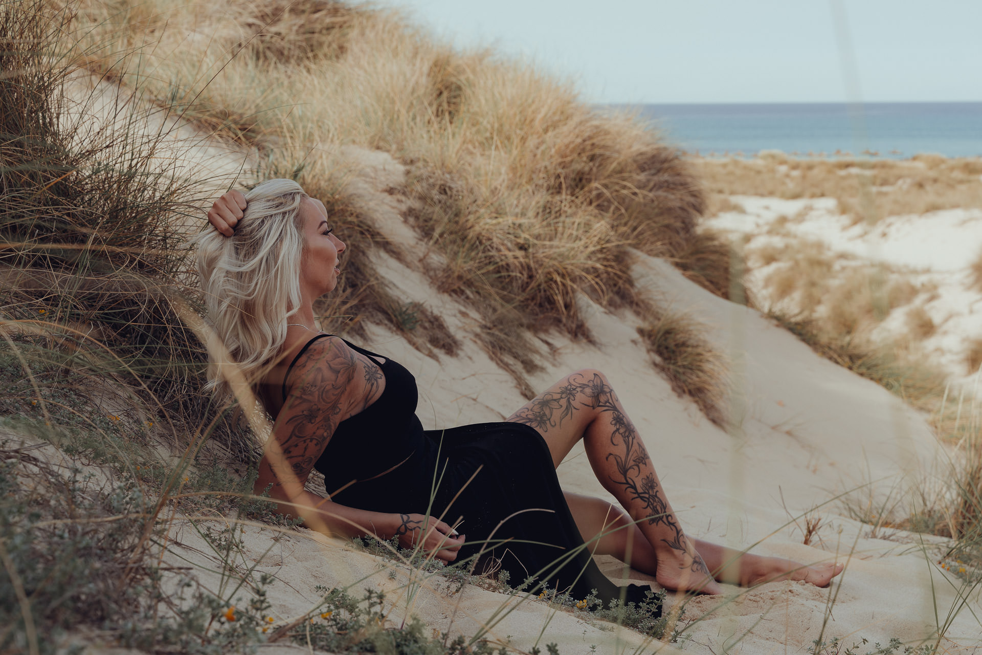 Tätowierte Frau lehnt gelassen im Dünengras mit weitem Blick über die Strände von Mallorca