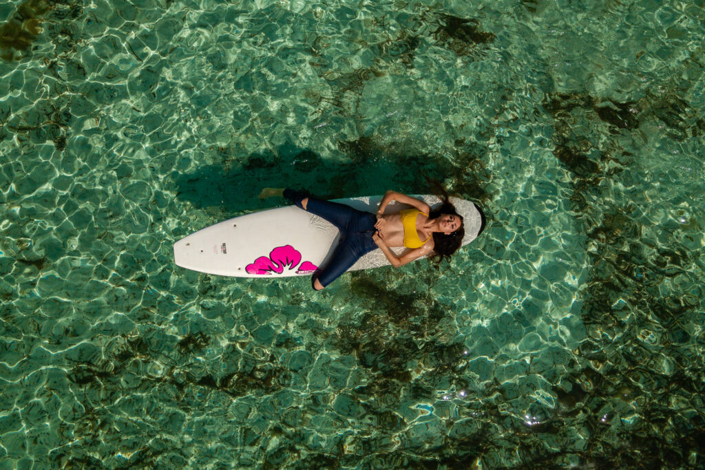 Luftbild eines Mallorca Model Shootings: Frau im gelben Bikini und Jeans relaxt auf dem Surfbrett im türkisfarbenen Wasser