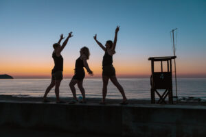 3 Mädchen tanzen auf der Mauer zum Strand von Cala Millor, iim Hintergrund kommt die Sonne aus dem Meer