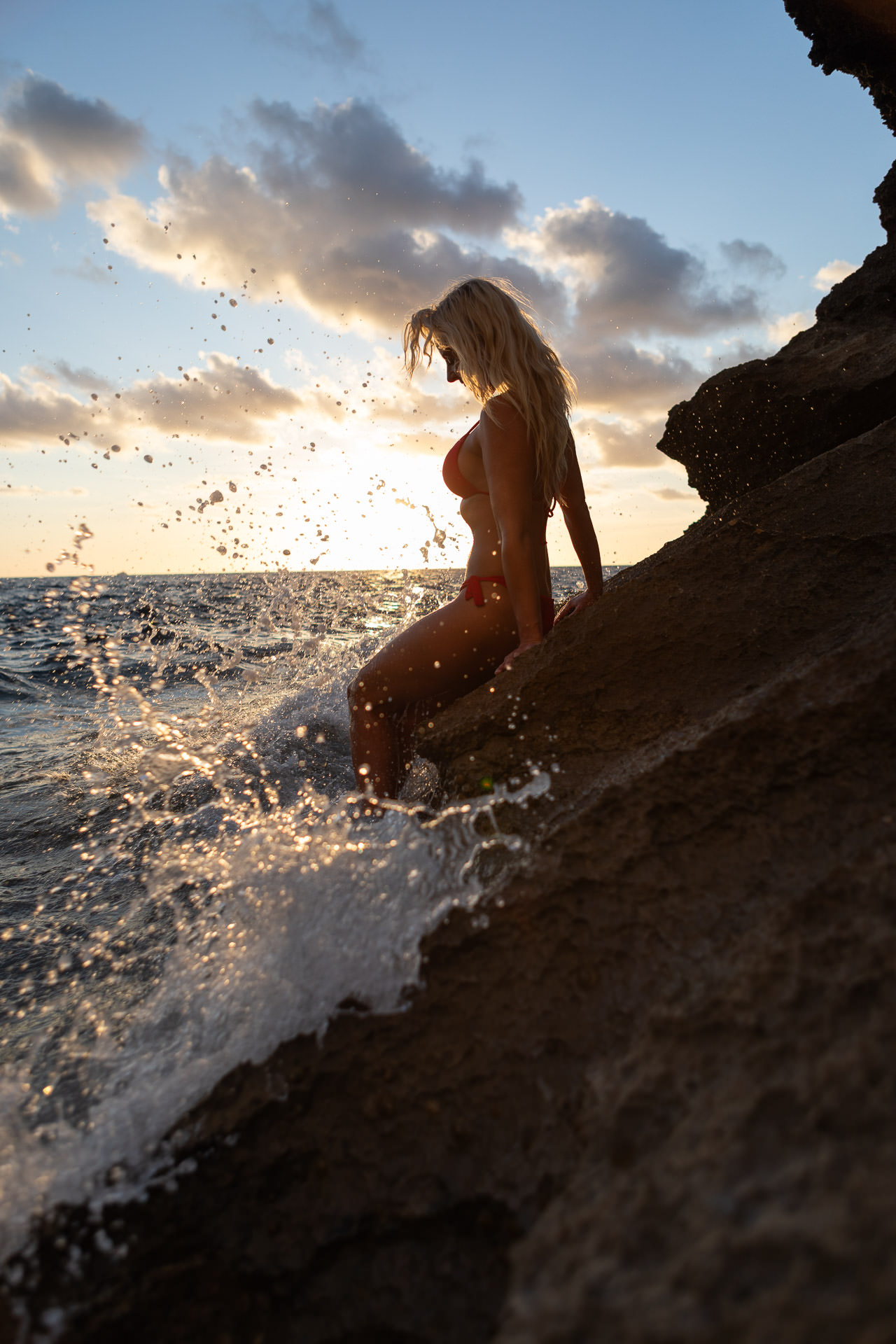 Mallorca Sonnenuntergang Shooting Model post an den Klippen in Mallorca, die wellen spritzen hoch und im Hintergrund ist der Sonnenuntergang zu sehen