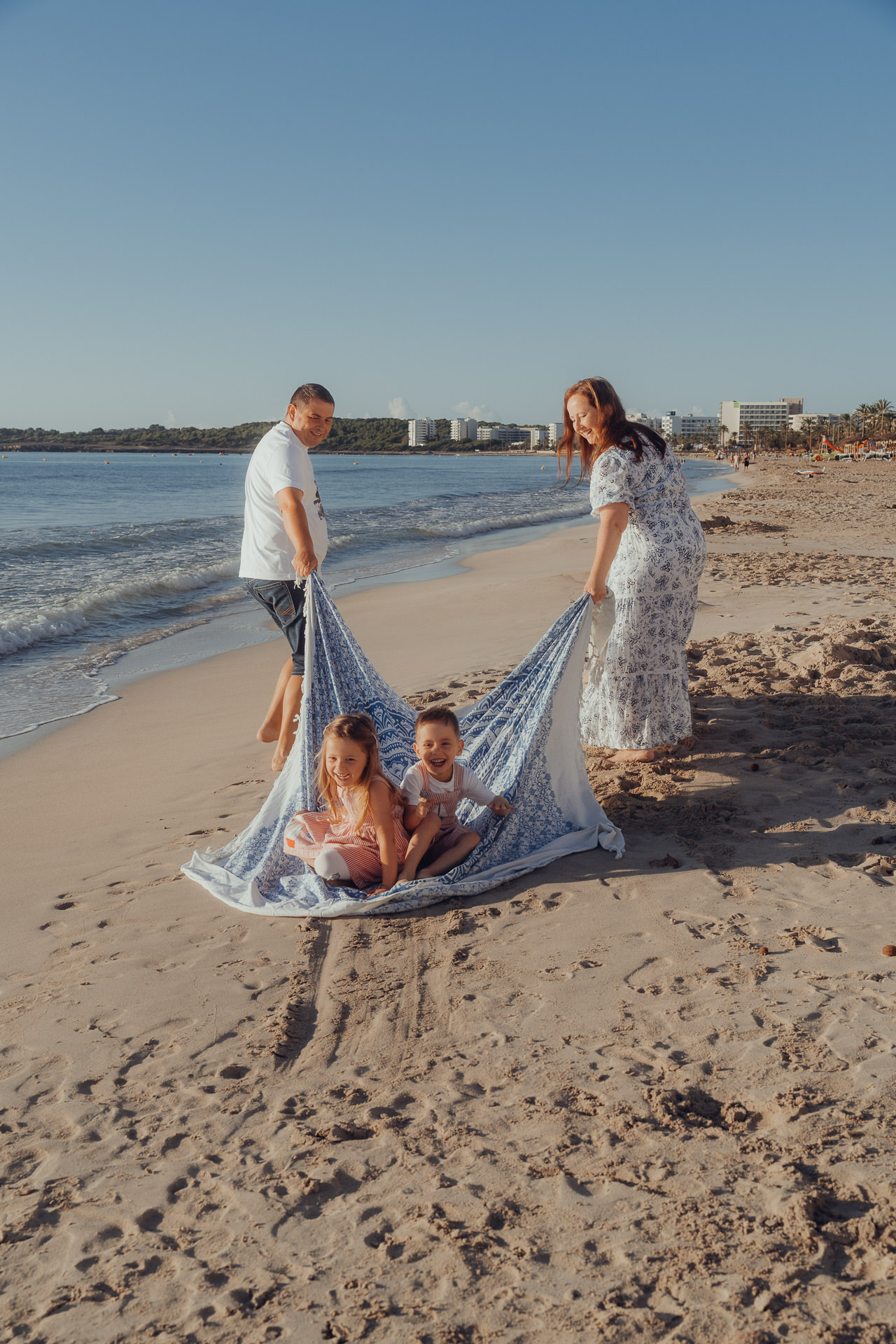 Mallorca Urlaubs Shooting Mutter und Vater ziehen 2 kleine Kinder auf einer großen Decke am Strand von Cala Millor