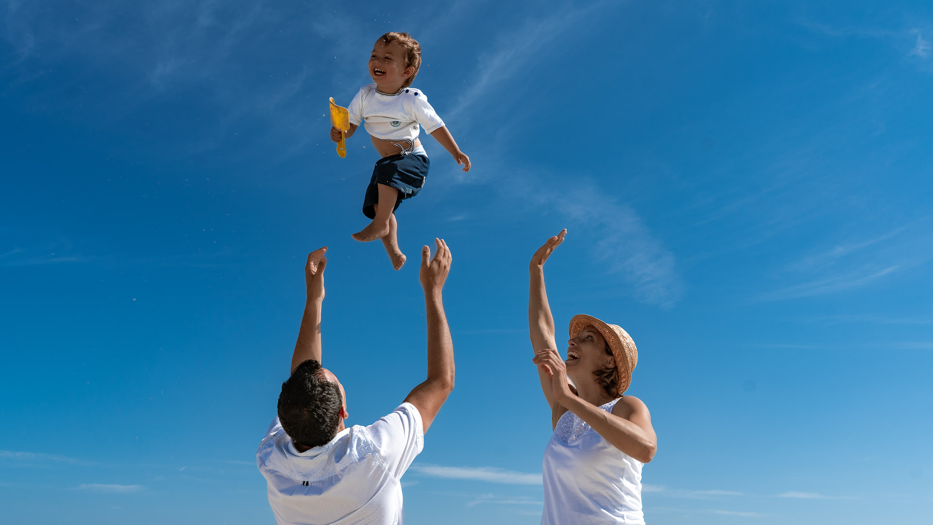 Ein kleiner junge wird von seinem Vater in die Luft geworfen und hat sehr viel Spaß dabei, die Mutter winkt dem Jungen zu.