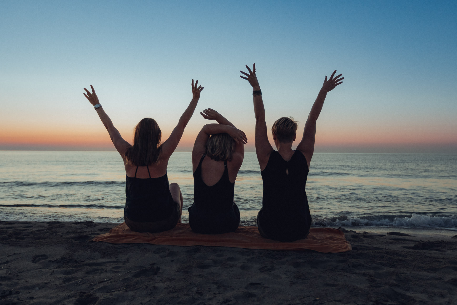 Mallorca Urlaubs Shooting Drei Freundinnen sitzen mit erhobenen Armen auf einem Handtuch am Strand von Mallorca und genießen den Sonnenaufgang.
