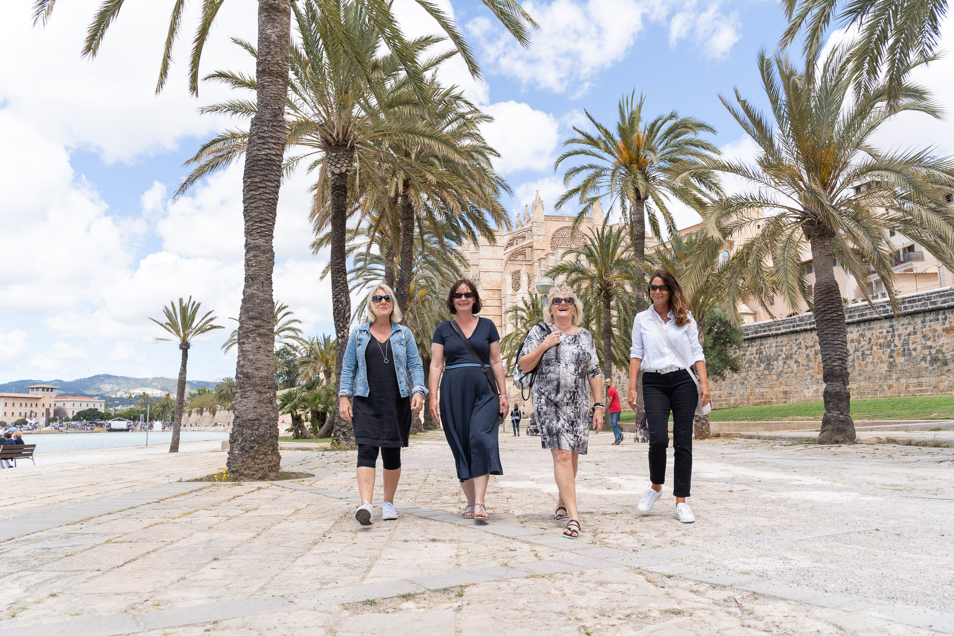 Freundinnen schlendern entspannt unter Palmen entlang, mit Blick auf die Kathedrale von Palma.