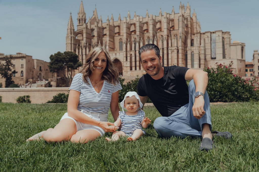 Junge Familie mit Baby sitzt lachend auf der Wiese vor der Kathedrale in Palma.