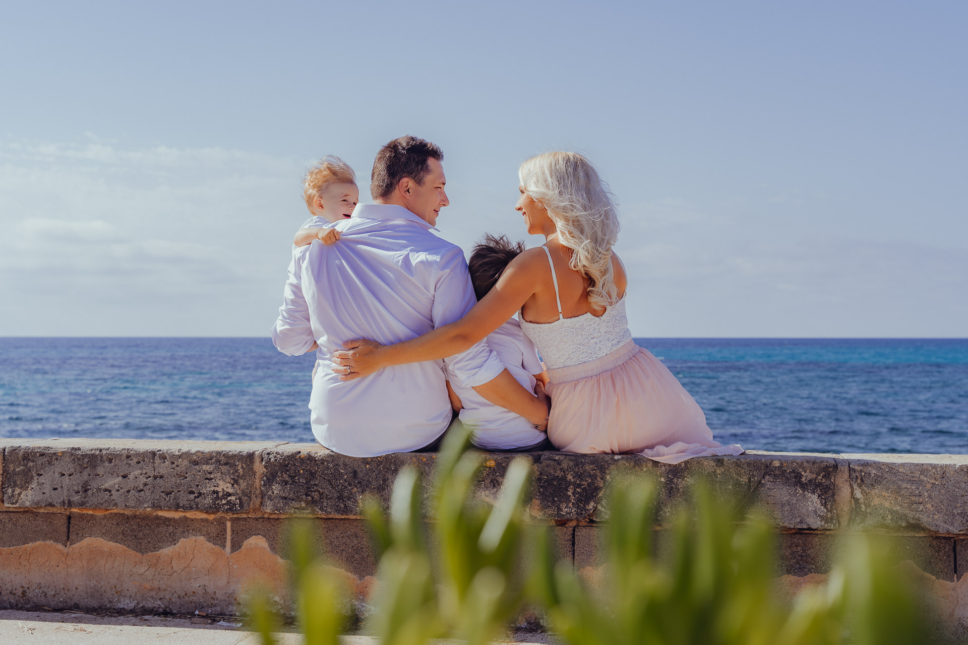 Familie sitzt beim Sa Coma Fotoshooting Arm in Arm auf einer Mauer am Meer und genießt die Aussicht auf das türkisfarbene Wasser