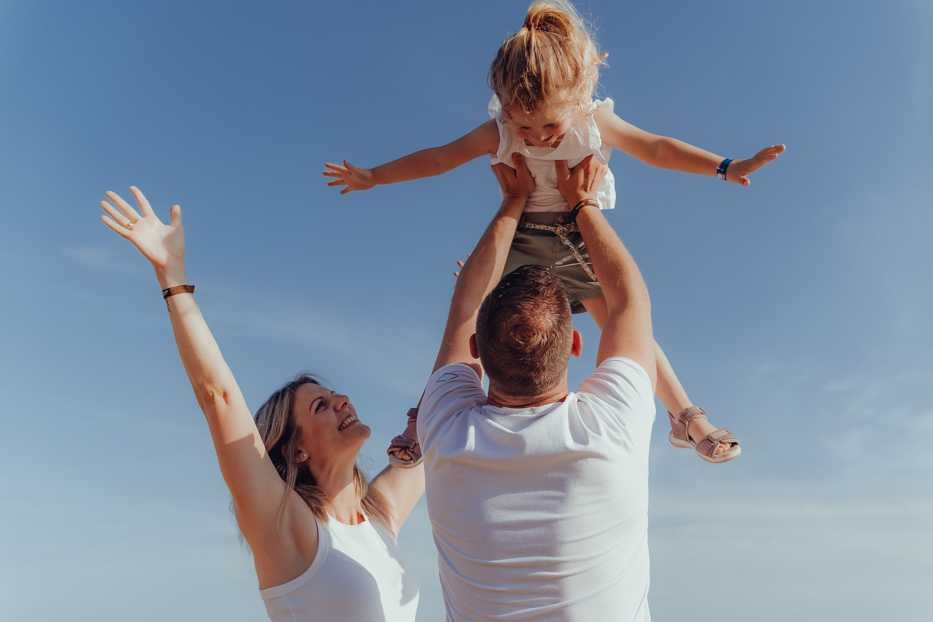 Eltern heben beim Sa Coma Fotoshooting lachend ihre Tochter in die Luft vor blauem Himmel am Strand von Sa Coma.