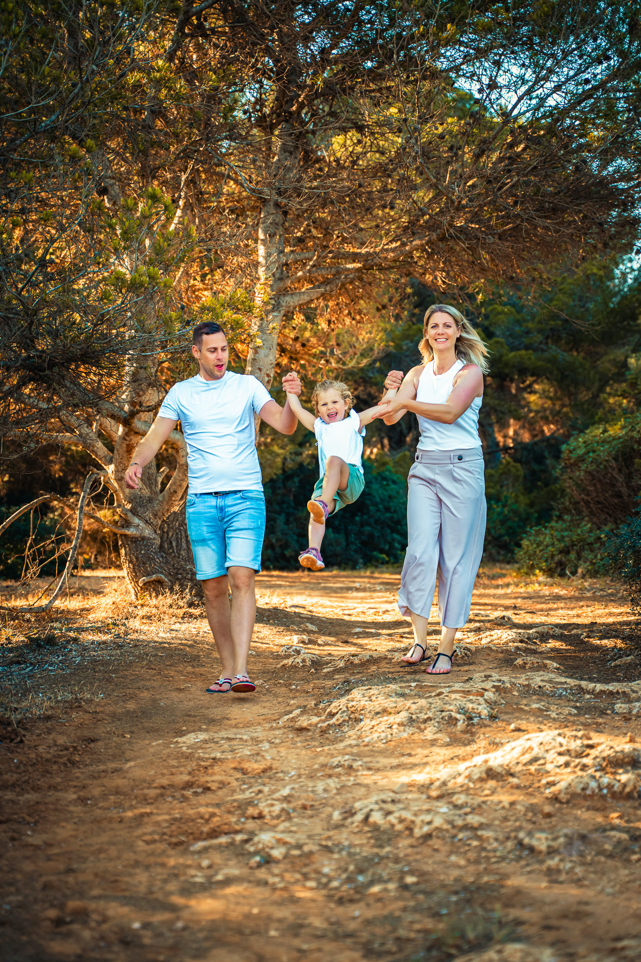 Glückliche Familie läuft beim Sa Coma Fotoshooting durch den Pinienwald und schwingt das Kind zwischen sich.