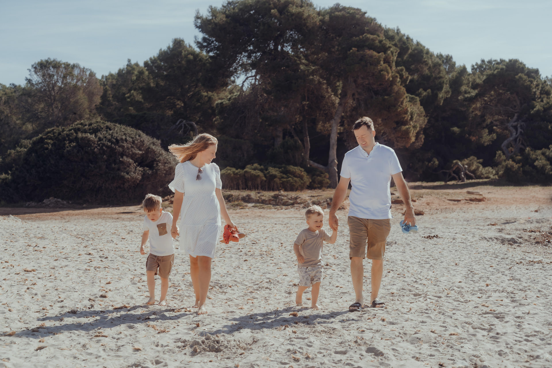 Familienspaziergang beim Sa Coma Fotoshooting: Eltern und zwei Kinder gehen gemeinsam barfuß über den weichen Sandstrand von Sa Coma