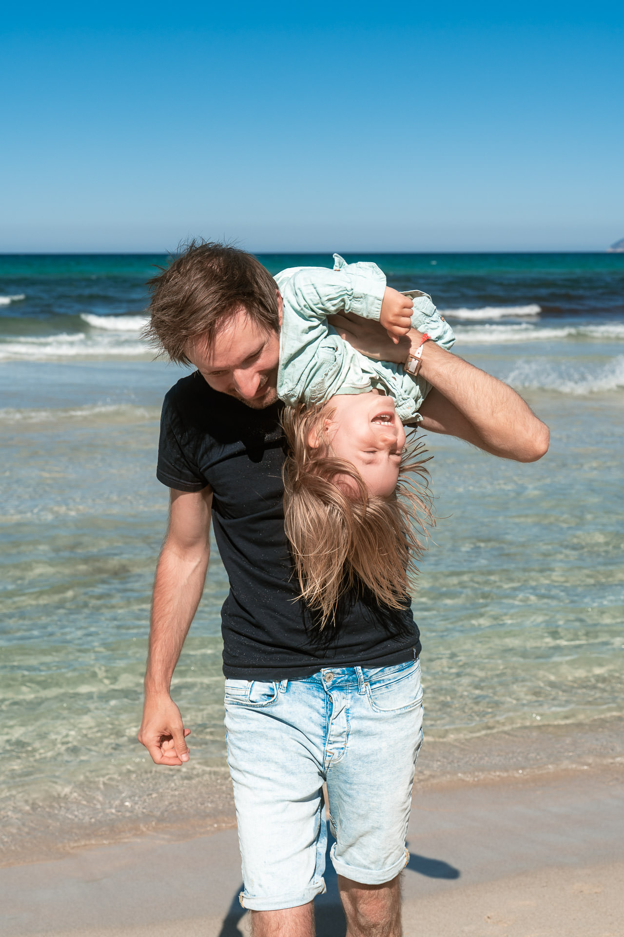 Vater trägt seine lachende Tochter beim Sa Coma Fotoshooting spielerisch über die Schulter am Wasser.