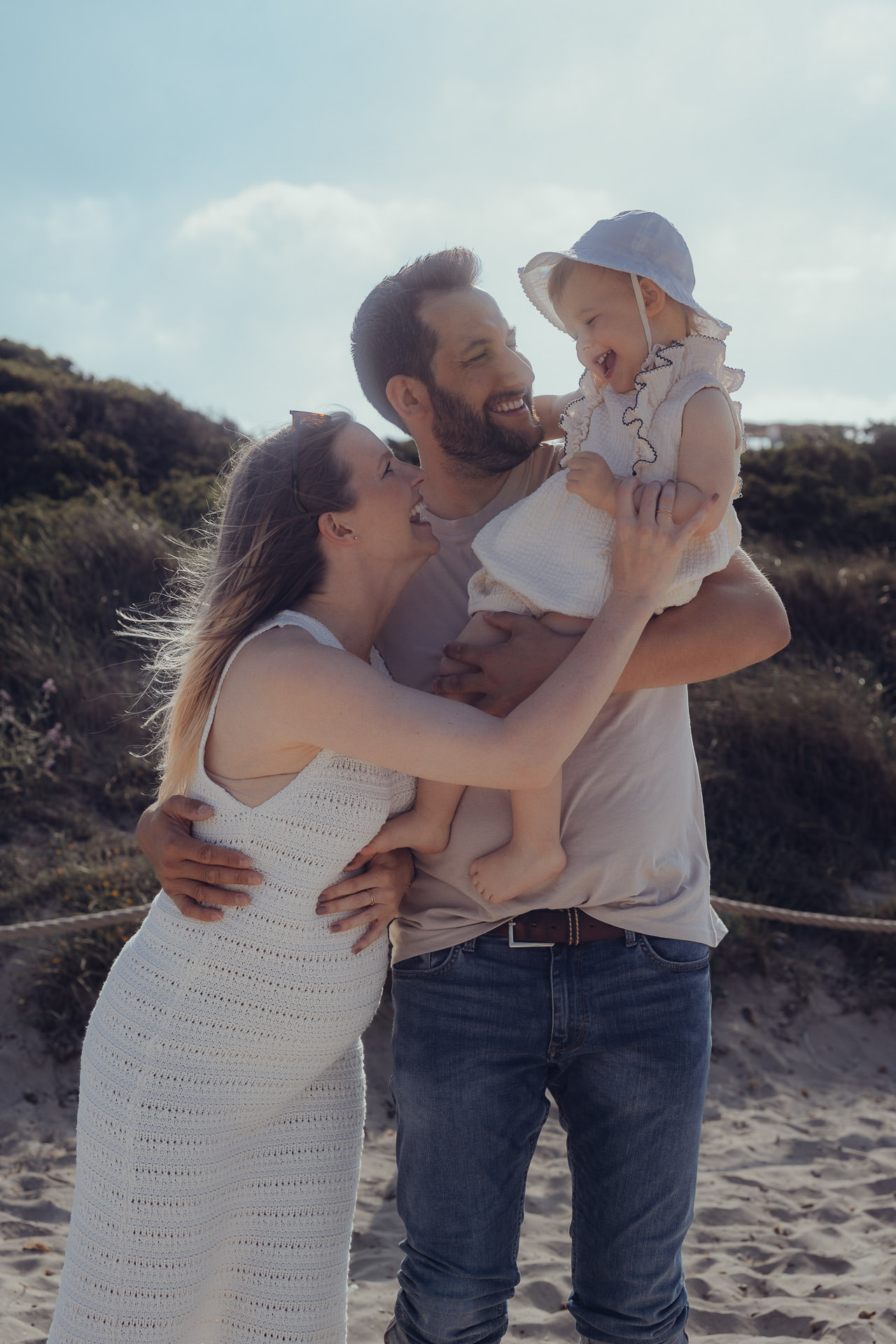 Inniger Moment beim Sa Coma Fotoshooting: Mutter, Vater und Kleinkind lachen und kuscheln am Dünenstrand von Sa Coma.