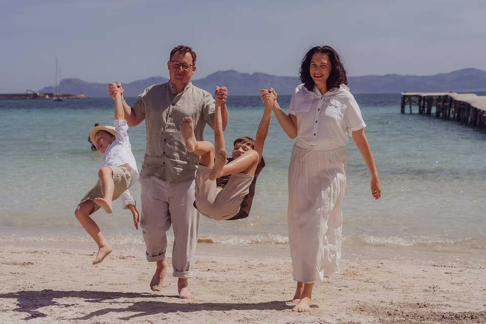 Familie mit zwei Kindern am Strand von Santa Ponça Mallorca, Eltern halten Kinder an den Händen beim Spielen am Meer, natürliche und lebendige Urlaubsfotos